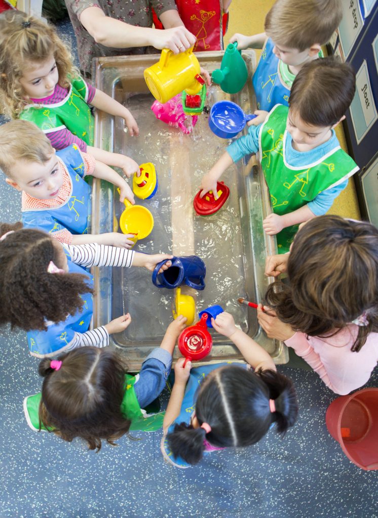 A number of children playing standing around a water trough playing with plastic toys.